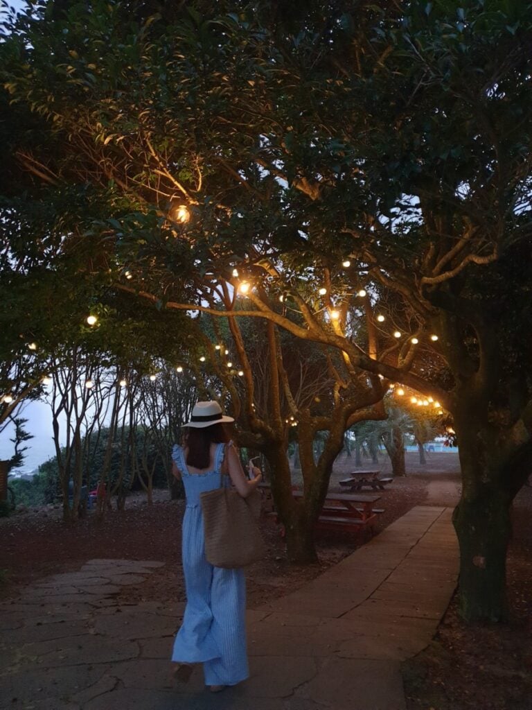 Rayoung walking among trees in Jeju with beautifully lit string lights on the trees.